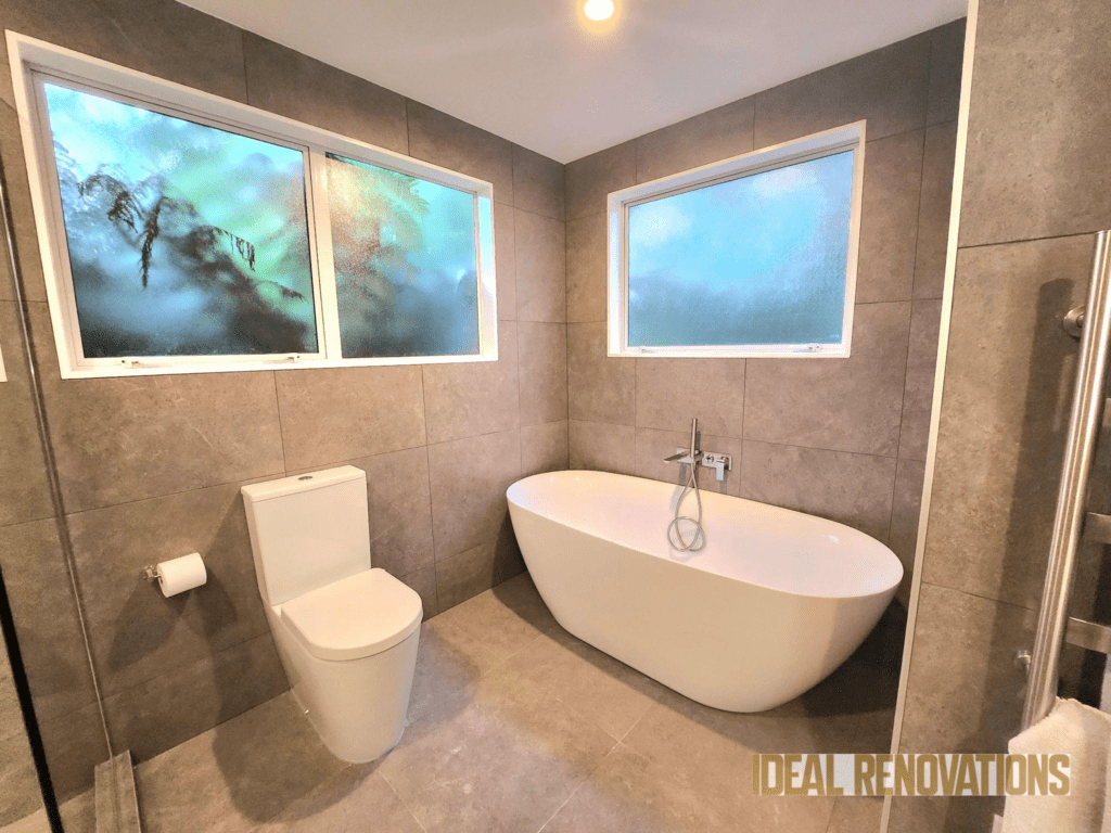 Modern bathroom renovation featuring a free-standing bathtub, wall-mounted toilet, and gray tiling, with frosted glass windows providing privacy